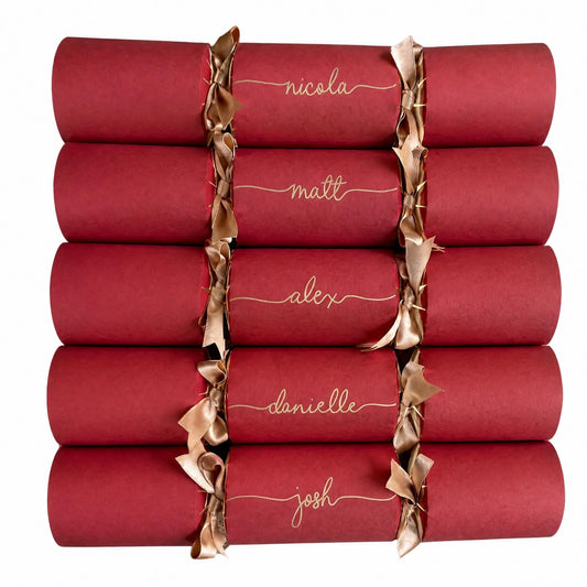 A collection of eight personalised red paper party Christmas crackers tied with double satin gold ribbon, each handwritten with a different name in gold ink, on a solid white background.