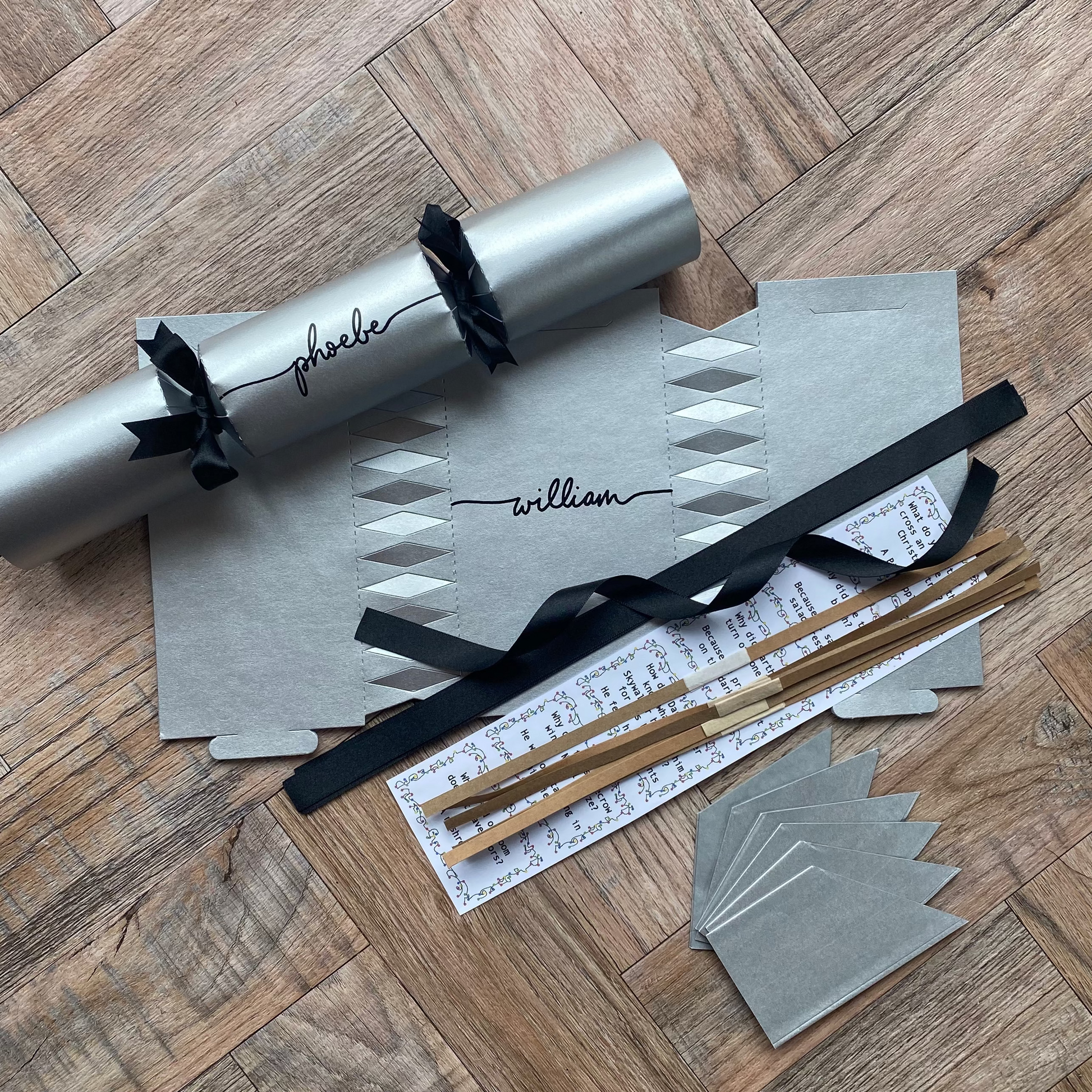 A flat-lay of a silver Christmas cracker with black ribbons, accompanied by matching silver party hats, black ribbons, cracker snaps, and a joke. 
a DIY Make Your Own Christmas Cracker Kit