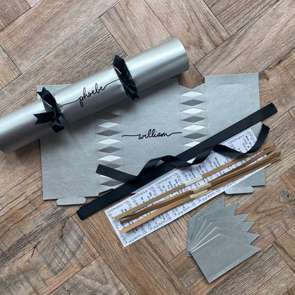 A flat-lay of a silver Christmas cracker with black ribbons, accompanied by matching silver party hats, black ribbons, cracker snaps, and a joke. 
a DIY Make Your Own Christmas Cracker Kit