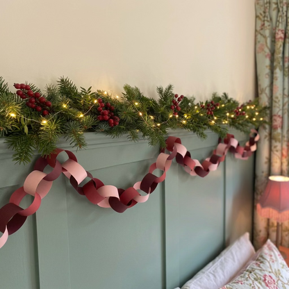 A paper chain garland with alternating shades of pink, burgundy, and deep plum draped across a teal paneled door next to a floral curtain.