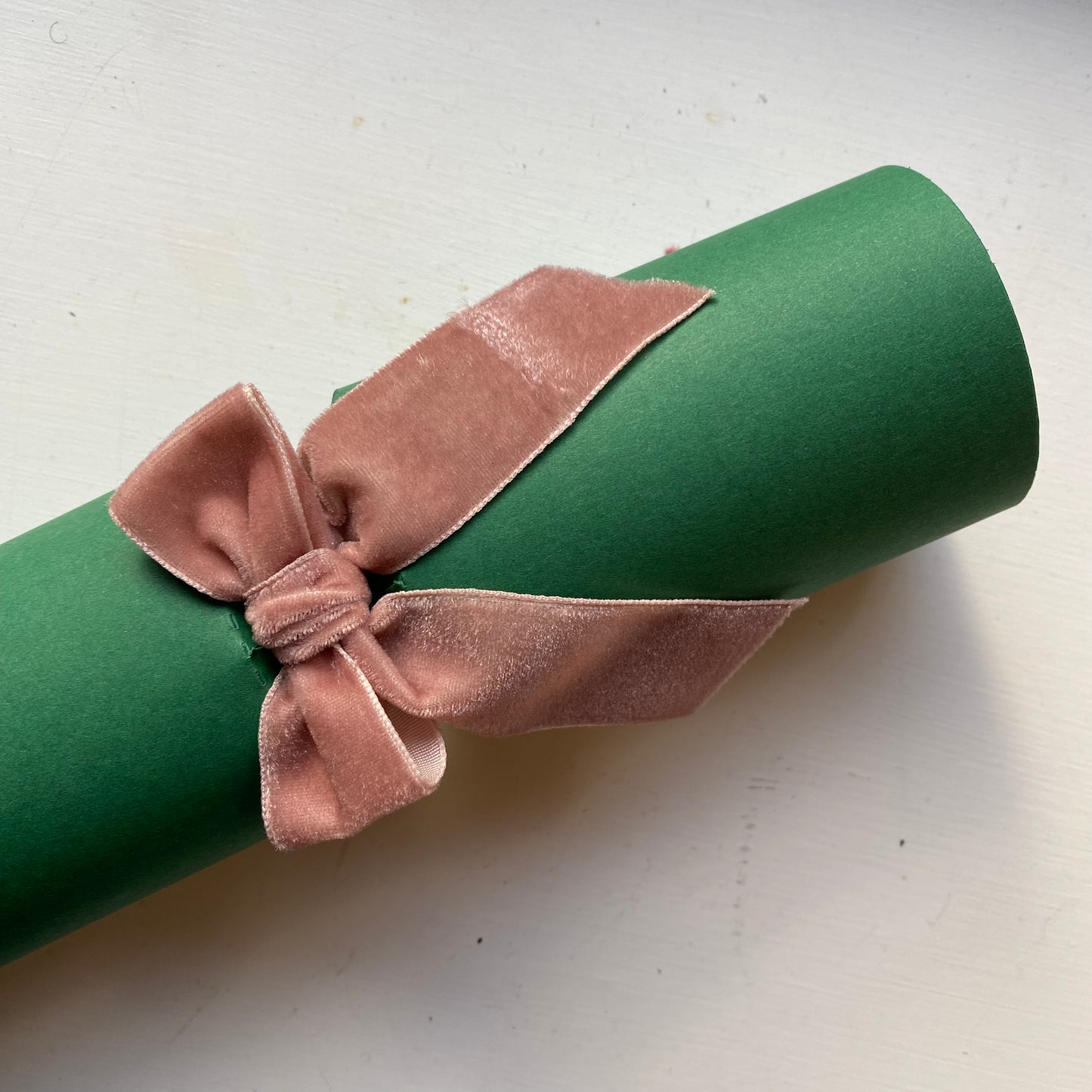 A green cylindrical gift tube or scroll wrapped with a dusty pink velvet ribbon tied in a bow, photographed on a light off-white surface.