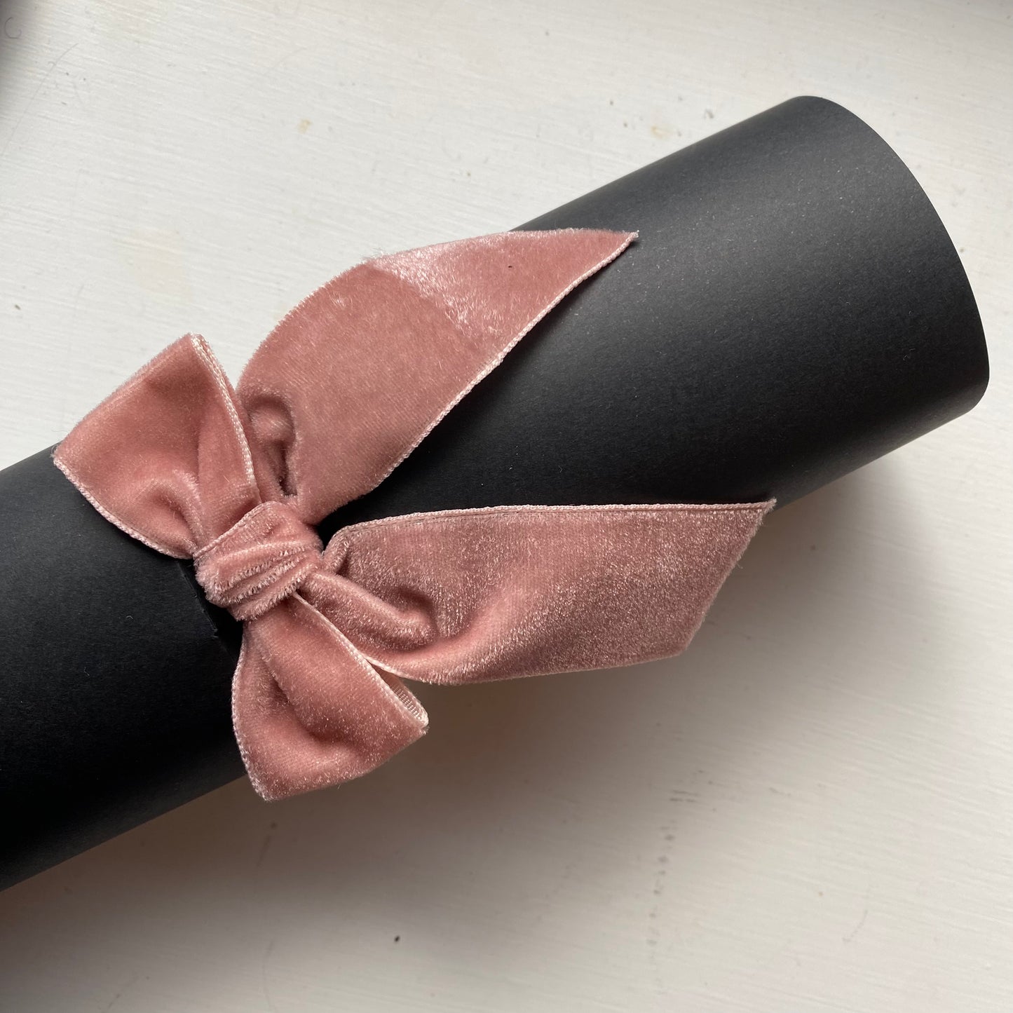 A dusty pink velvet bow tied around a black cylindrical display stand on a light off-white surface.
