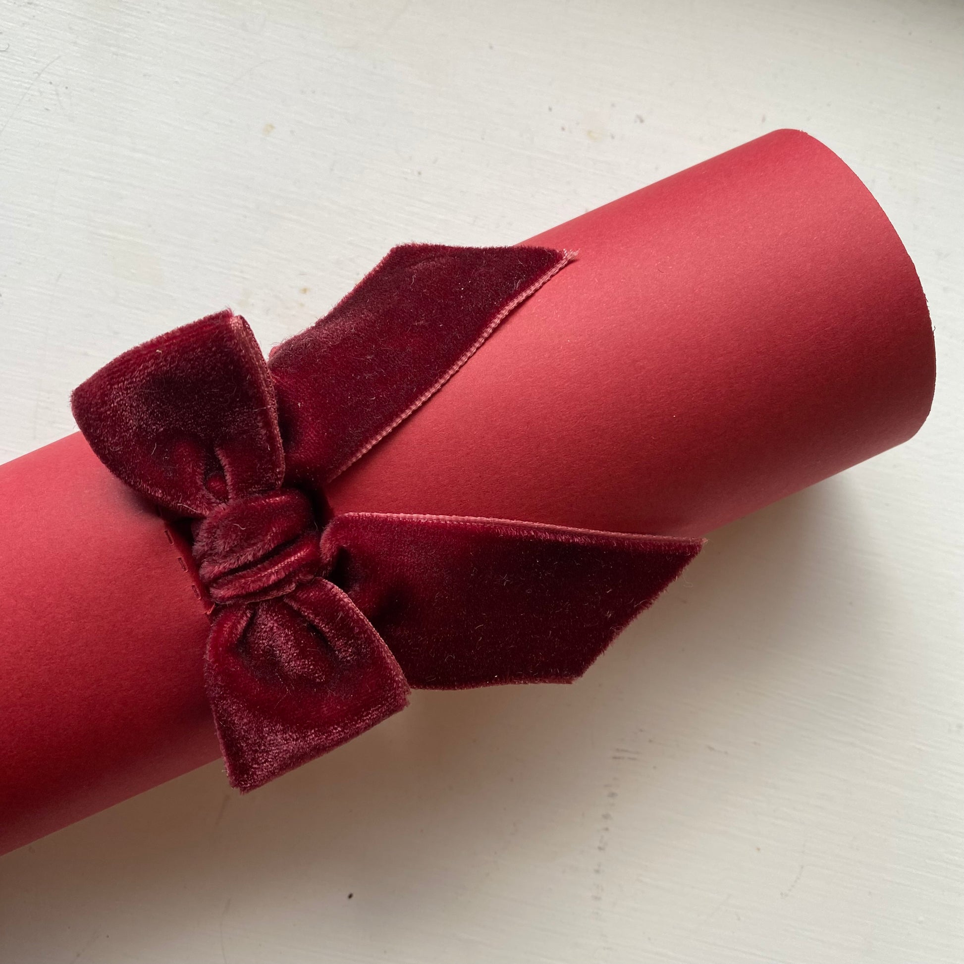 A rolled red CHRISTMAS CRACKER tied with a matching red velvet bow, photographed on a light off-white background.